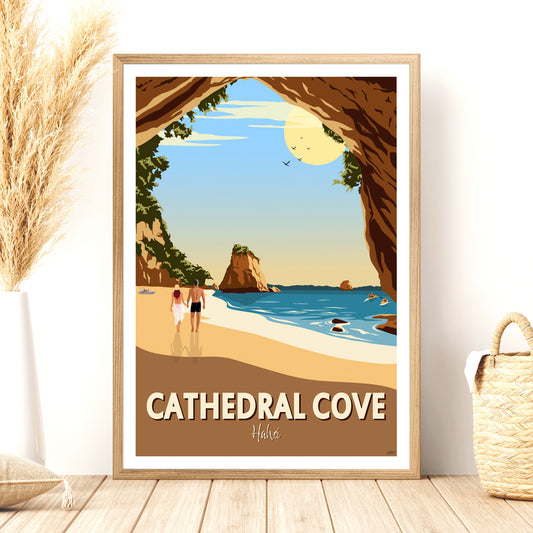 A framed travel poster depicting Cathedral Cove, showing two people walking on the beach with rock formations and the cove in the background. The frame is placed on a wooden floor with a straw hat and dried plants to the side.