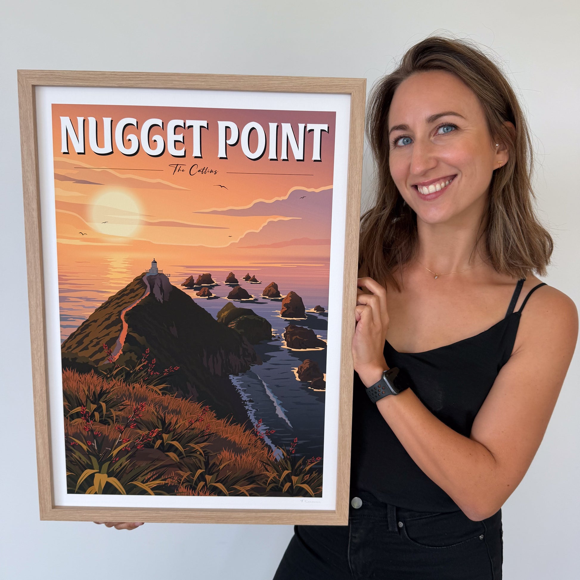 Woman holding a framed artwork titled 'Nugget Point' with a scenic depiction of a sunset over water.