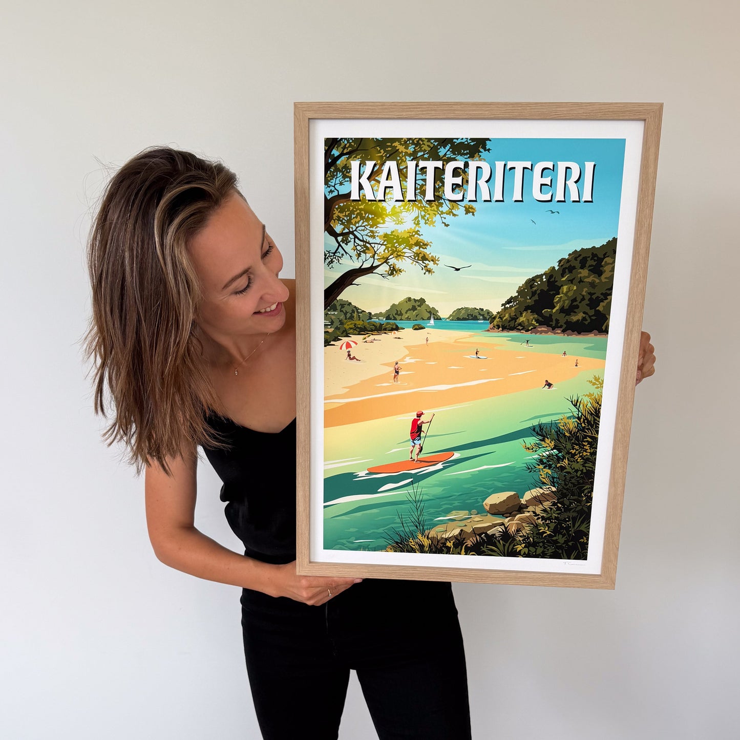 Person holding a framed artwork of Kaiteriteri with a scenic beach and people on a paddleboard.