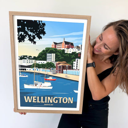 A woman in a black top and black pants holds up a framed travel poster of Wellington, Ontario, Canada. The poster features a vintage-style illustration of a harbor with boats, a green hillside town, and a red-roofed building under a blue sky.